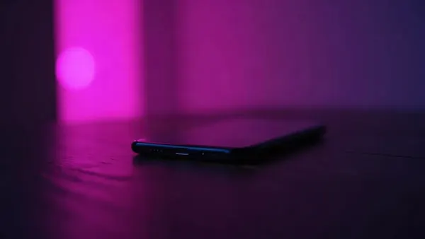 Smartphone auf einem dunklen Holztisch, umgeben von violetten und rosa Reflexen (Illustration zum Artikel)